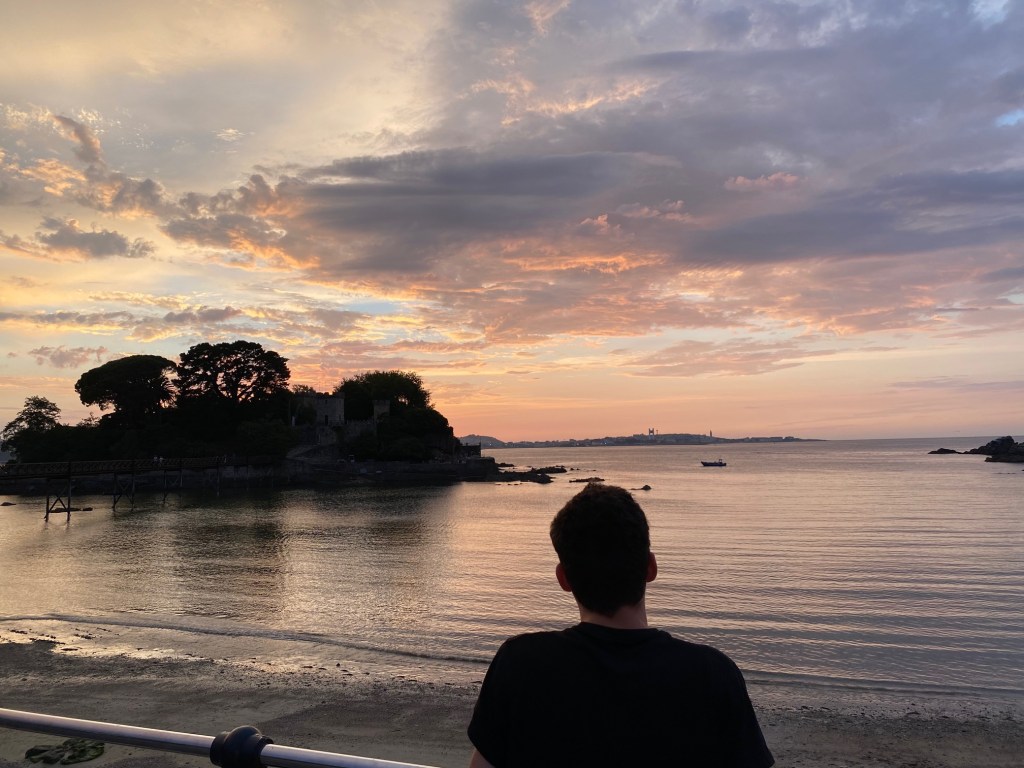 puesta de sol sobre el mar con el cielo de color rojizo. A la derecha se recorta la silueta de un chico que está de espaldas y a la izquierda la de una isla