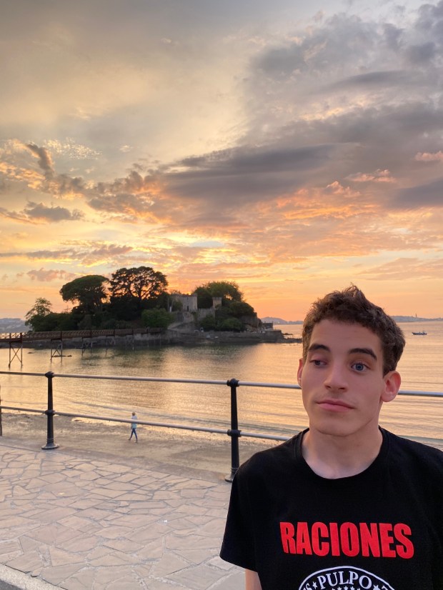 Adolescente con una camisa negra con letras rojas que imita la clásica de "Los Ramones" pero que lo cambia por "Raciones". Al fondo se ve una isla con un castillo y un cielo rojo sobre el mar que anuncia la puesta del sol.