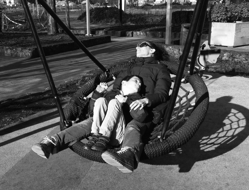 Foto en blanco y negro de una madre y su hijo meciéndose en un columpio nido. El niño tiene unos 7-8 años y está tumbado sobre su madre, que lo abraza y protege con sus brazos. Llevan ropa de invierno. Están tumbados de frente y el sol les da de lleno en el rostro. El niño tiene la cabeza girada hacia su izquierda y los ojos cerrados para protegerse y la madre se intuye que también, aunque lleva gafas de sol.
