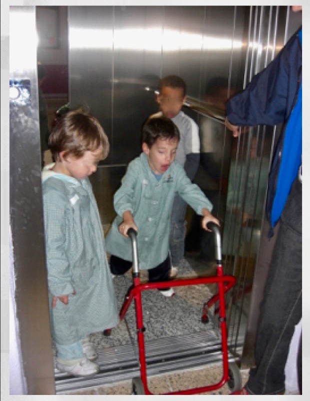 Foto de tres niños pequeños saliendo de un ascensor. Los tres llevan batas de cuadritos verdes y uno de ellos utiliza un andador.