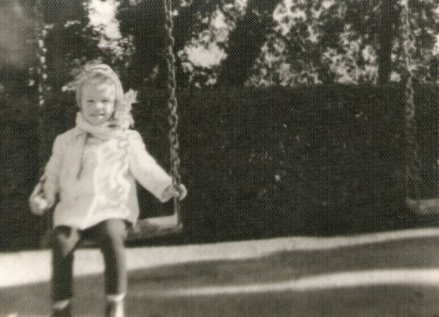 Foto en blanco y negro de una niña de unos tres años sentada en un columpio. Lleva un pantalón negro, un chaqueta clara y aparece abrigada con gorro y bufanda.