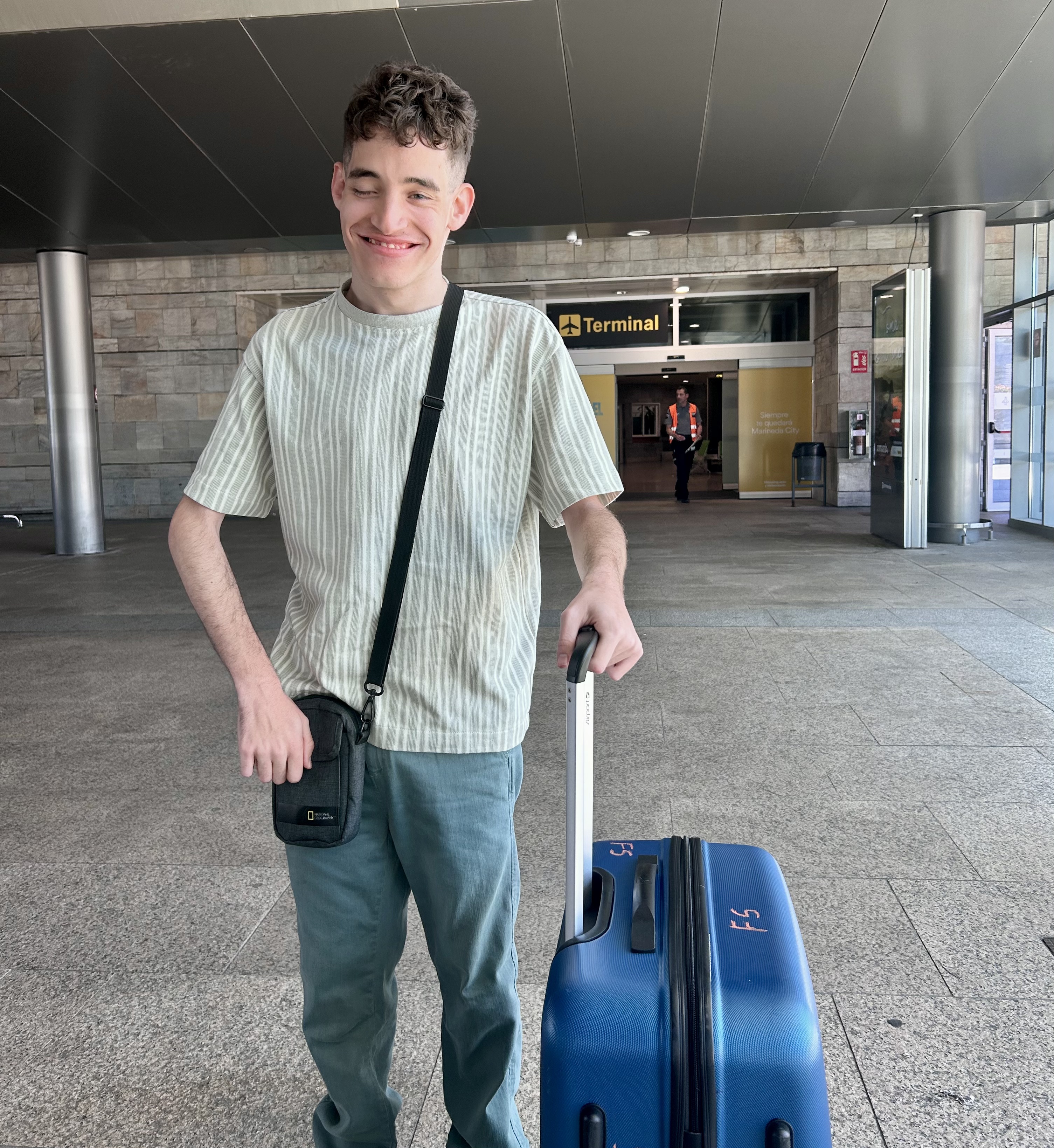 Imagen de Antón sonriente y sosteniendo el asa de una maleta de ruedas delante de la puerta de entrada al aeropuerto